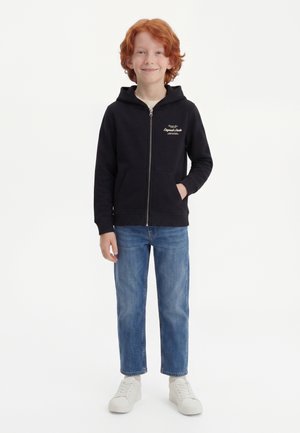 Enfant roux debout, portant un sweat à capuche noir zippé, un jean bleu et des baskets blanches, souriant à la caméra sur un fond blanc.