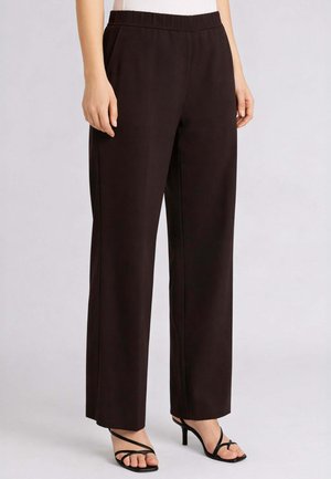 Donna con pantaloni neri a gamba larga e vita alta abbinati a sandali neri con tacchi e cinturini, in piedi davanti a uno sfondo semplice.