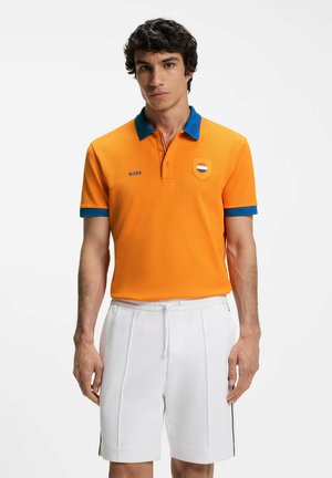 Uomo che indossa una polo arancione con colletto e bordo blu, pantaloncini bianchi con coulisse, in piedi davanti a uno sfondo bianco uniforme.