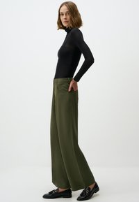 Maglia nera a maniche lunghe con colletto a dolcevita abbinata a pantaloni larghi verde oliva, con tasche a toppa e una textura liscia. Mocassini neri completano l'outfit.