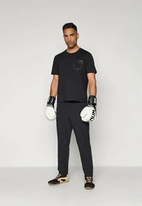 Muž oblečený v čiernom športovom oblečení a bielych boxerských rukaviciach proti jednoduchému svetlému pozadiu.