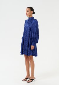 Sininen silkkinen mini mekko korkealla kauluksella, pitkät hihat ja nappimansetit; siinä on pehmeä rakenne ja hienovarainen kuviointi koko ajan.