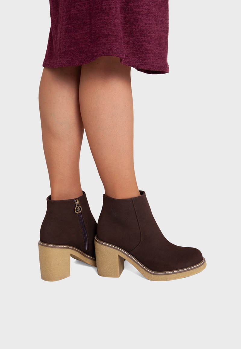 Jambes portant une jupe bordeaux et des bottines marron foncé avec des talons beiges épais et des fermetures éclair latérales sur un fond uni.