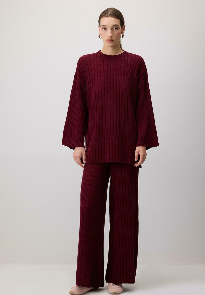 Kobieta ubrana w prążkowany, luźny czerwony sweter i pasujące szerokie spodnie, stojąca na jednolitym szarym tle.