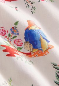 Tkanina we wzory przedstawiająca ilustrację niedźwiedzia w niebieskim płaszczu i pomarańczowym kapeluszu, niosącego teczkę, otoczonego kwiatowymi wzorami w różnych kolorach.