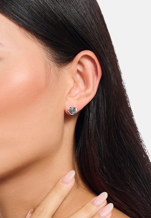 Silberne Ohrringe in Form einer rose, mit einem strukturierten Design. Am Ohrläppchen getragen, ergänzt durch glattes, dunkles Haar und natürliche Nägel.