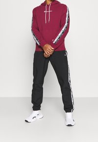 Hoodie bordeaux avec logo blanc et cordons, pantalon noir avec rayures blanches, et chaussures de sport blanches. Tenue décontractée et sportive.