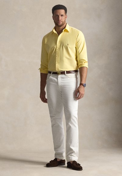 Bărbat în picioare purtând cămașă galbenă cu nasturi, pantaloni albi, curea maro, mocasini maro cu ciucuri și ceas de mână pe fundal neutru.