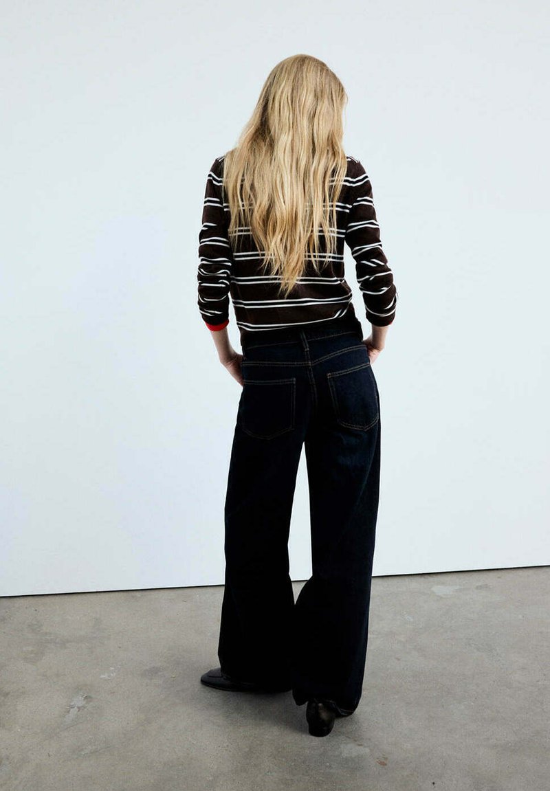 Femme blonde aux longs cheveux portant un pull rayé noir et blanc et un jean large foncé, debout face à un mur blanc.
