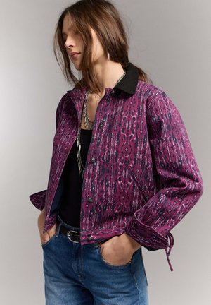 Mujer con cabello castaño que lleva una chaqueta morada con estampado y cuello negro, y vaqueros azules, con las manos en los bolsillos, sobre un fondo claro y liso.