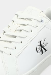Baltos odinės sportbačiai su plokščiomis baltomis raištelėmis, subtiliomis siūlėmis ir iškilnia juoda Calvin Klein logo šone. Lygus paviršius, minimalistiškas dizainas.