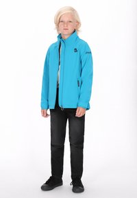 Modra softshell jakna z visokim ovratnikom, sprednjim zadrgo, črnimi akcenti, oblečena čez sivo srajco, v kombinaciji s črnimi hlačami in čevlji.