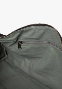 Das Innere der Stofftasche verfügt über ein glattes olivgrünes Futter, eine kleine Reißverschlusstasche und einen dunklen Rand. Der Reißverschluss hat eine bronzeartige Oberfläche.