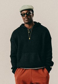 Άνδρας με μαύρη πλεκτή κουκούλα, κόκκινες παντελόνες, πράσινο καπέλο τύπου flat, μαύρα γυαλιά ηλίου και χρυσές αλυσίδες στέκεται με τα χέρια στις τσέπες.