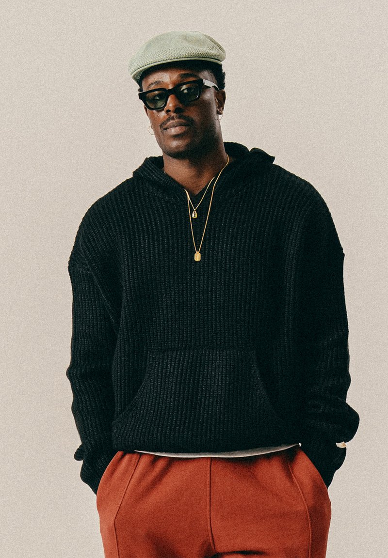 Άνδρας με μαύρη πλεκτή κουκούλα, κόκκινες παντελόνες, πράσινο καπέλο τύπου flat, μαύρα γυαλιά ηλίου και χρυσές αλυσίδες στέκεται με τα χέρια στις τσέπες.