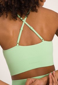 Light green ribbed crop top with thin, crossed straps and adjustable hardware detailing. Smooth texture, fitted design, and minimal branding.