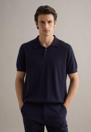 Joven con cabello oscuro que lleva una camiseta polo azul marino de manga corta y pantalones texturizados azul marino, de pie con las manos en los bolsillos contra un fondo neutro.