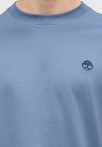 Ανοιχτό μπλε μπλουζάκι με στρογγυλή λαιμόκοψη και μικρό σκούρο μπλε κεντητό λογότυπο δέντρου στο στήθος.