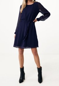 Vestido azul marino con mangas largas transparentes, cintura ceñida con lazo y patrón de espiga. Largo hasta la rodilla con un corte recto. Lleva botas negras hasta el tobillo.