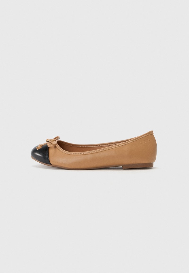 Chaussure de ballet plate beige pour femme avec un embout arrondi noir et un petit nœud à l'avant, présentée sur un fond clair uni.