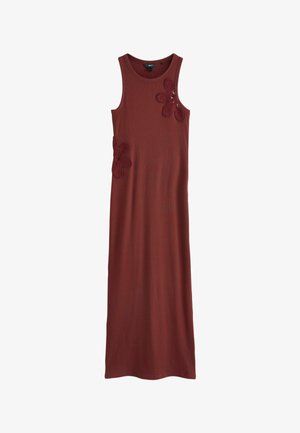 Femme en robe maxi sans manches couleur rouille avec des détails floraux, debout à l'intérieur près d'un canapé avec des coussins blancs et une grande fenêtre.