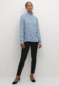 Blusa a maniche lunghe azzurro chiaro con motivo geometrico nero, colletto alto e arricciato, abbinata a pantaloni neri aderenti e tacchi neri.