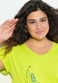 Camiseta verde lima de cuello en v con mangas cortas, que presenta una impresión gráfica de un velero y el texto "LA VIDA ESTÁ AQUÍ". Textura suave.