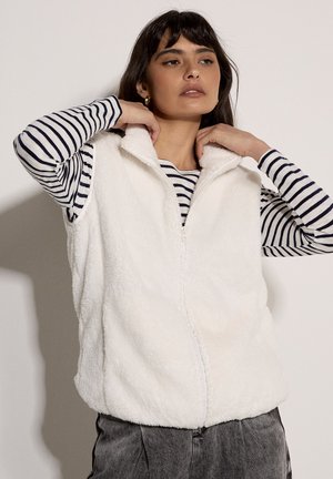 Weißes, flauschiges ärmelloses Weste mit hohem Kragen und Reißverschluss vorne. Wird über einem gestreiften Langarmshirt getragen. Weiche Textur und lässiges Design.