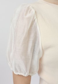 Krēmkrāsas tops ar caurspīdīgu pūkainu piedurkni, kurā plecā un aprocē ir savilkts elastīgais audums, redzams uz personas augšējās rokas un pleca.