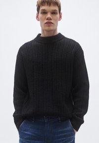 Mustast kudumist kampsun, millel on kõrge soonikuga kaelus ja pikad varrukad, millel on tekstuurse mustriga ja lõdva istuvusega, kantud siniste teksapükste kohal.