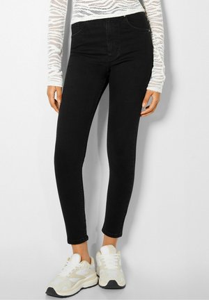 Vrouw met zwarte skinny jeans, wit kanten shirt met lange mouwen en witte sneakers, staand tegen een effen achtergrond.