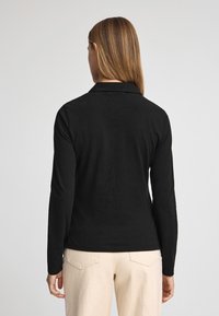 Camisa polo negra de manga larga hecha de tejido suave, con cuello y diseño ajustado, combinada con pantalones beiges, mostrando una vista trasera.