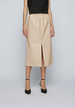 Jupe midi en simili cuir beige avec une taille élastique, dotée d'une fente sur le devant et d'une texture lisse, assortie à des talons noirs et un haut noir.