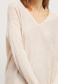 Jasnobeżowy sweter z dekoltem w serek o luźnym kroju. Teksturowana dzianina i opuszczone ramiona nadają swobodny wygląd.