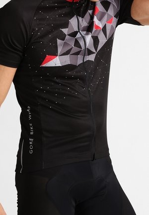 Nahaufnahme einer Person, die ein schwarzes Radtrikot mit geometrischen Mustern in Grau und Rot sowie gepolsterte schwarze Radhosen trägt.