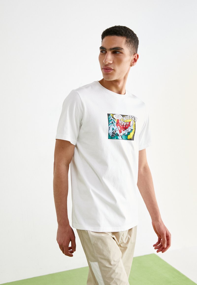 Camiseta blanca de algodón con un estampado gráfico colorido en la parte delantera. Corte casual con mangas cortas, combinada con pantalones color beige claro.