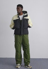 Padded jacket s čierno-krémovým farebným blokovaním, so zipsom a kapucňou; skombinovaná so zelenými cargo nohavicami a viacfarebnými teniskami.