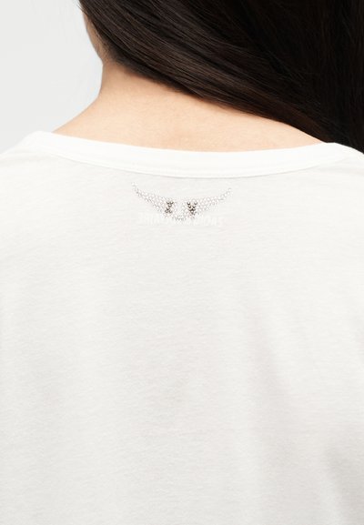 Κοντινό πλάνο του άνω μέρους της πλάτης ενός ατόμου με μακριά σκουρόχρωμα μαλλιά που φοράει άσπρο πουκάμισο με μικρό ασημένιο σχέδιο φτερού από στρας κοντά στη λαιμόκοψη.