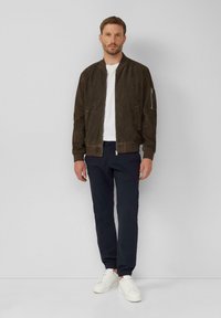 Dunkelbraune Wildleder-Bomberjacke mit gerippten Bündchen und Kragen, vorne mit Reißverschluss und seitlichen Taschen, getragen über einem weißen Hemd und navyblauen Hosen.
