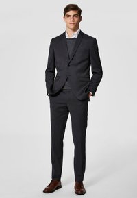 Mørk grå dress med skreddersydd passform, med notched lapel, en-knaps lukking og rette bukser. Bæres over en lys genser og skjorte. Brune sko.