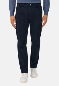 Tumed sinine puuvillane püksid sirge lõikega, millel on kaks esitaskut ja diskreetsed õmblused, koos valgete spordijalatsitega.