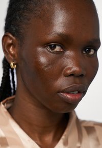 Nærbilde av et ansikt med mørk hudtone, med glatt tekstur og subtil glans. Gull øredobber og bluse i nøytrale toner med diagonale striper.