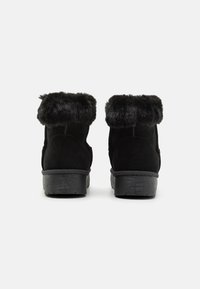 Botas negras hasta el tobillo con un suave collar de pelo sintético, material de ante, tacón grueso negro y suela texturizada. La vista trasera muestra detalles de costura.