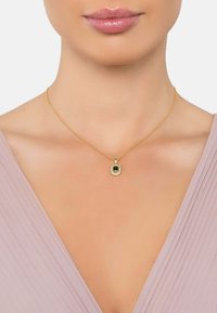 Collana d'oro con un pendente rettangolare di pietra verde, circondato da gemme trasparenti. Maglie della catena sottili, indossata su uno sfondo di tessuto morbido e rosa.