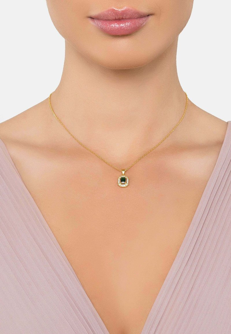 Collana d'oro con un pendente rettangolare di pietra verde, circondato da gemme trasparenti. Maglie della catena sottili, indossata su uno sfondo di tessuto morbido e rosa.
