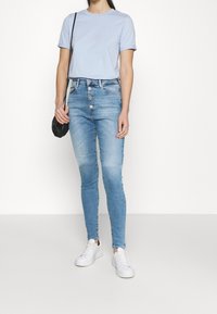 Camiseta de algodón azul clara combinada con unos vaqueros de tiro alto y corte ajustado en un tono azul desgastado, con cinco botones. Zapatillas blancas completan el atuendo.