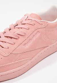 Gros plan sur une sneaker Reebok en daim rose avec lacets, détails de couture et une semelle texturée sur un fond blanc.