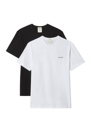 Hombre de pie con las manos detrás de la espalda, vistiendo una camiseta blanca de Calvin Klein, pantalones negros, cinturón negro y zapatos negros sobre un fondo gris liso.