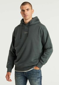 Felpa grigia scura con vestibilità rilassata, dotata di cappuccio con lacci e logo discreto sul petto. Indossata sopra una maglietta grigia chiara e jeans blu.