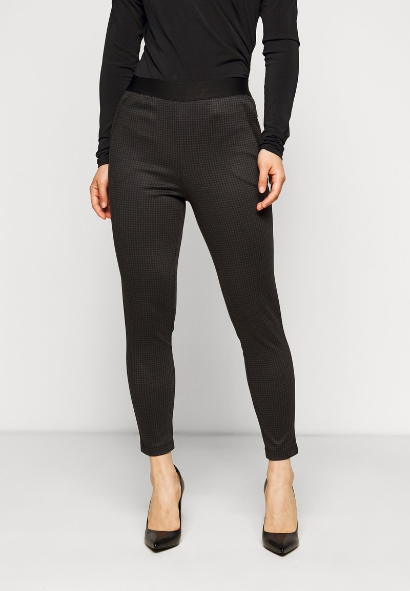 Femme portant un pantalon à carreaux noir et gris ajusté, un haut à manches longues noir et des chaussures à talons noirs, debout devant un fond uni.